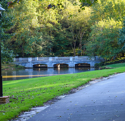 a stone bridge over the lake surrounded by green grass and lush green and autumn colored trees with still silky brown lake water at Lenox Park in Brookhaven - cheap car insurance in Georgia.