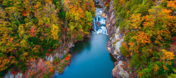 An aerial view shows a narrow waterfall dropping into a calm blue pool inside a rocky gorge, surrounded by dense forest in peak fall colors—reds, oranges, and greens—in Forest Park, Georgia. Cheap car insurance in Georgia.
