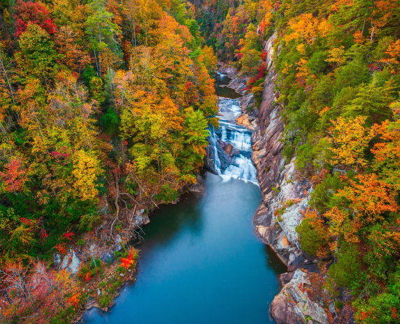An aerial view shows a narrow waterfall dropping into a calm blue pool inside a rocky gorge, surrounded by dense forest in peak fall colors—reds, oranges, and greens—in Forest Park, Georgia. Cheap car insurance in Georgia.
