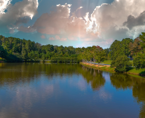 A stunning panoramic shot of the still green lake surrounded by lush green and autumn colored trees with powerful clouds at sunset at Duncan Park in Fairburn Georgia USA.