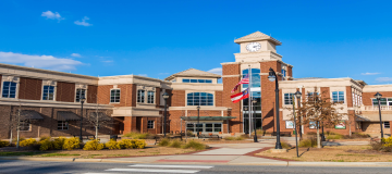Street view of Lilburn City Hall, showing outside sitting area, main entrance doors and windows, and pole with flags of USA and Georgia, under fair skies.