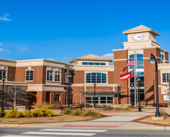 Street view of Lilburn City Hall, showing outside sitting area, main entrance doors and windows, and pole with flags of USA and Georgia, under fair skies.